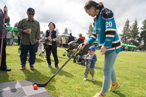 woman in sharks jersey on grass with hockey stick about to shoot puck.