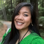 Person smiling with long black hair, wearing a green top, with redwoods behind her. 